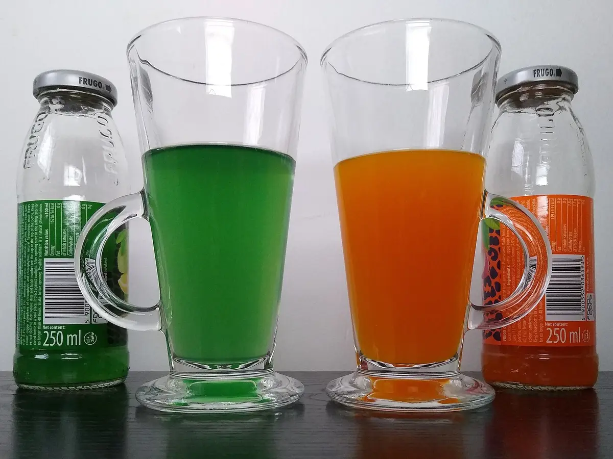 Zwei Gläser mit grüner und oranger Limonade neben den passenden Glasflaschen
