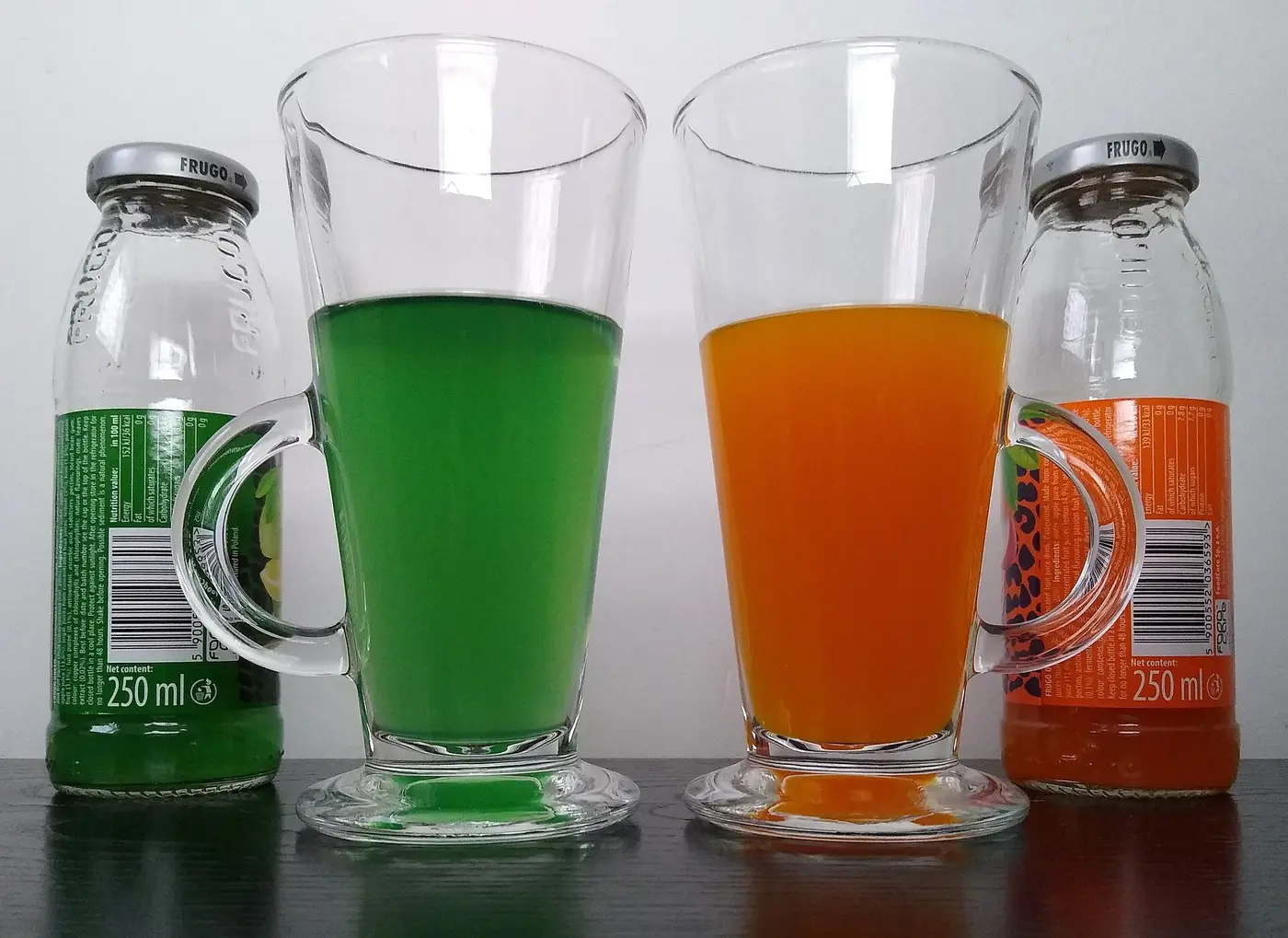 Two glasses of green and orange soft drink next to their glass bottles